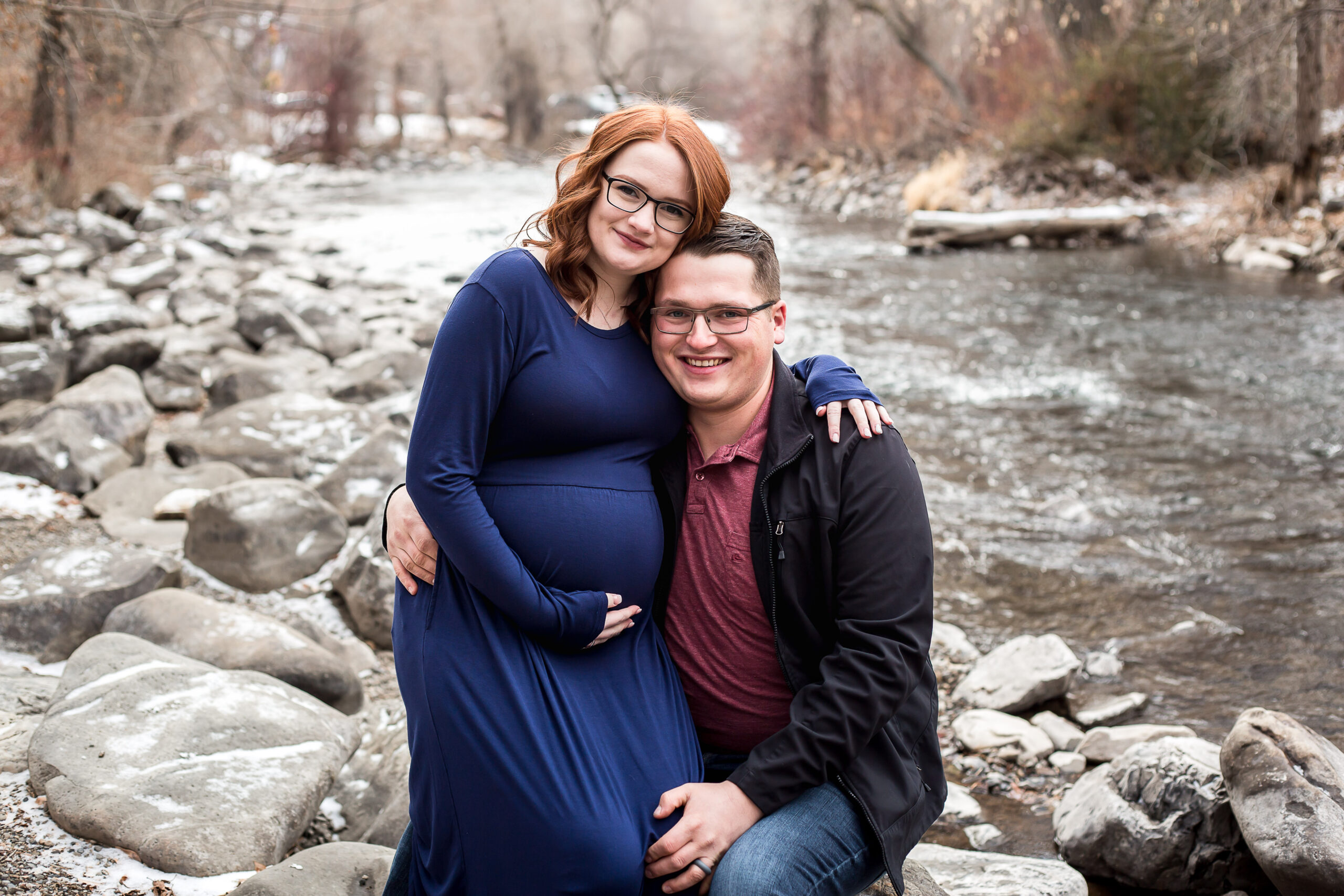 A couple sit by a river posing for a photo during their maternity session. They are anxiously awaiting the birth of their first child, a daughter.